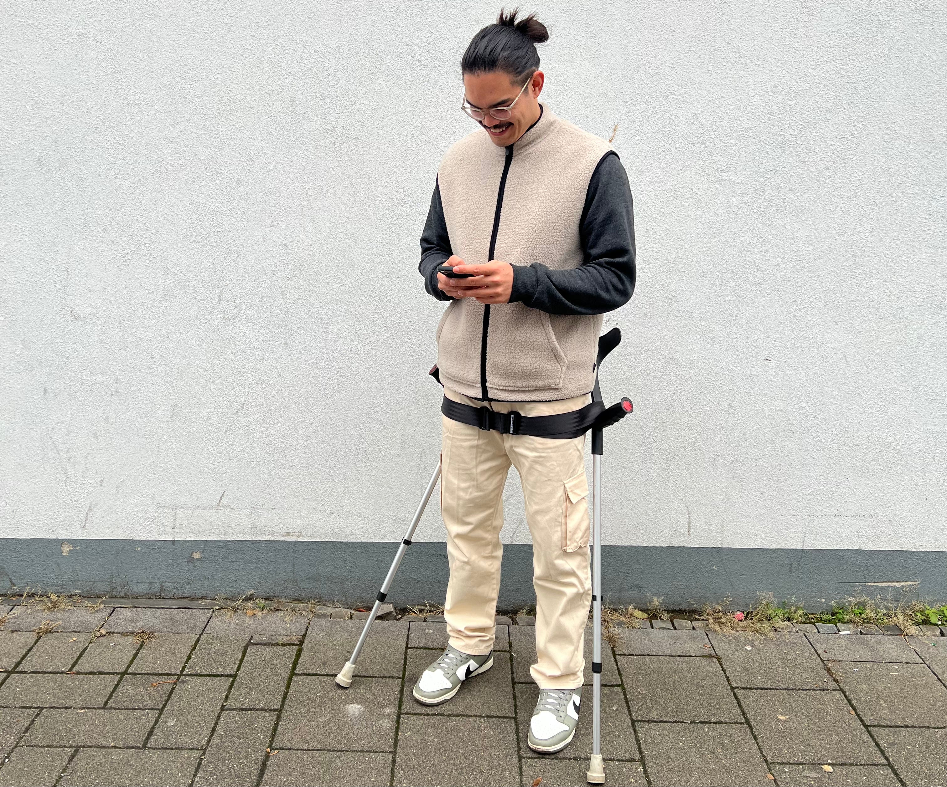 Mann hat seine Krücken an der Hüfte abgestellt und ist am Handy.